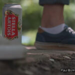 A cold beer can placed next to a person's foot, set against the backdrop of a sunlit park