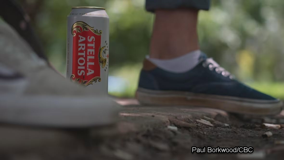 A cold beer can placed next to a person's foot, set against the backdrop of a sunlit park